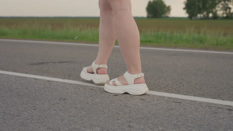 leg view of fair skin woman walking along asphalt road marked by white line beside green verge under soft summer sunlight showing casual stride and smooth motion in rural outdoor setting