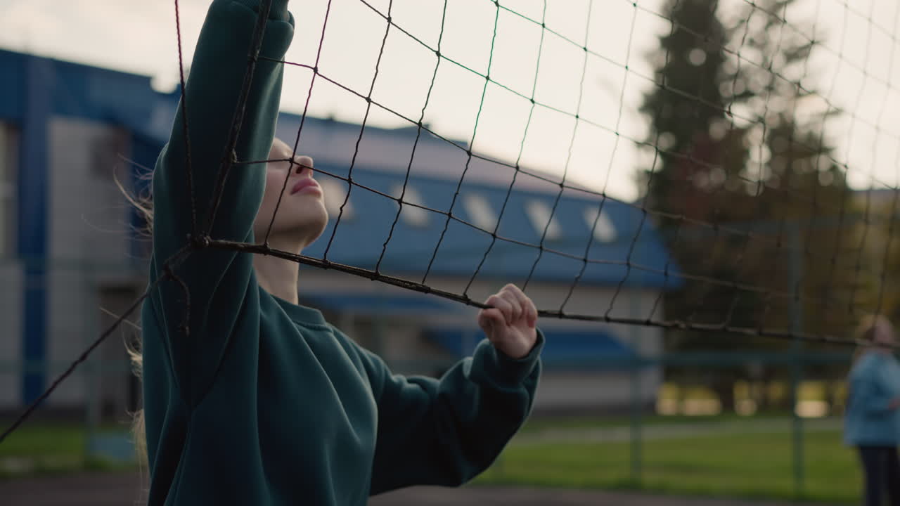 Close up of sportswoman in green sweater holding and shaking volleyball net with blurry background of someone playing volleyball, outdoor sports training session with sunlight and sports facility