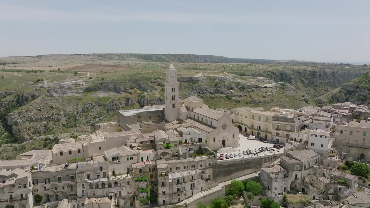 Aerial drone view of ancient hilltop town Matera Italy showing stone buildings, bell tower, narrow streets, and surrounding natural landscape under clear sky in daylight