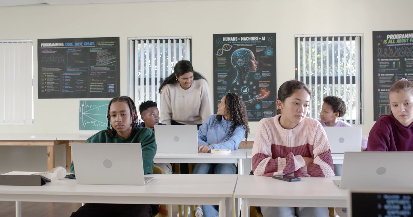 Adolescenti di diverse etnie lavorano sui laptop in classe mentre un insegnante indiano cammina e si china per aiutare un compagno.
