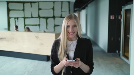 young formal woman using smartphone in office. stylish blond woman in jacket standing in contemporary office hall and surfing smartphone with smile