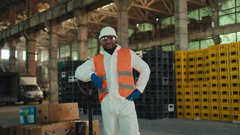 Portrait-of-a-confident-man-with-Black-skin-in-a-white-protective-uniform-and-an-orange-vest-who-stands-in-safety-glasses-near-large-boxes-for-sorted-waste-at-a-plant-for-processing-and-sorting-garbage-and-waste