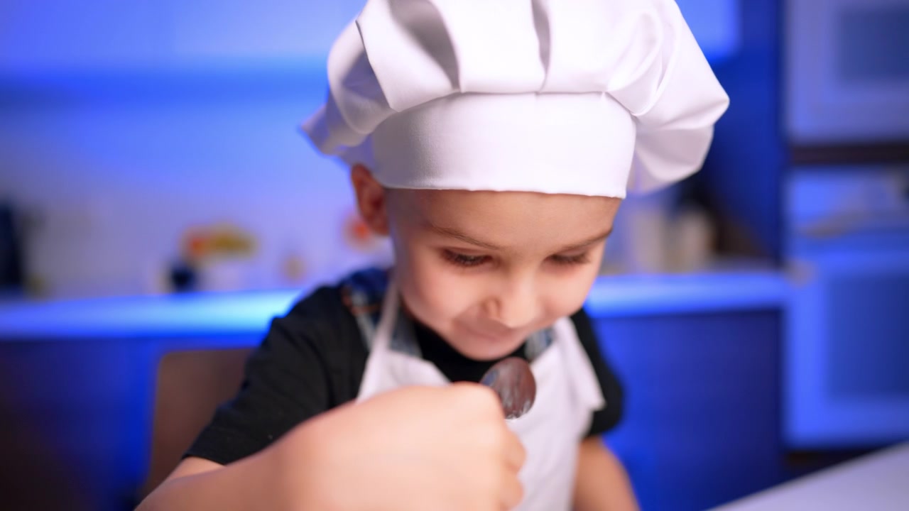 Primo piano di un bambino in uniforme da chef che mangia con un cucchiaio. Un bambino assaggia della crema bianca da un cucchiaio su uno sfondo sfocato con illuminazione blu in una cucina.