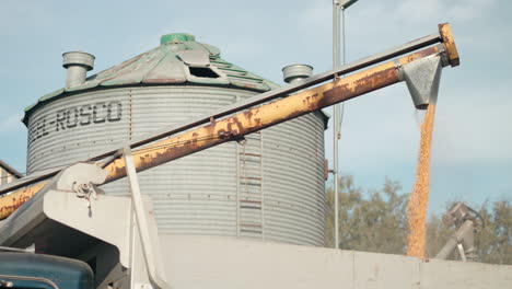 Maiskörner,-Die-Aus-Einem-Mähdrescher-Mit-Riesigem-Silo-Im-Hintergrund-Strömen