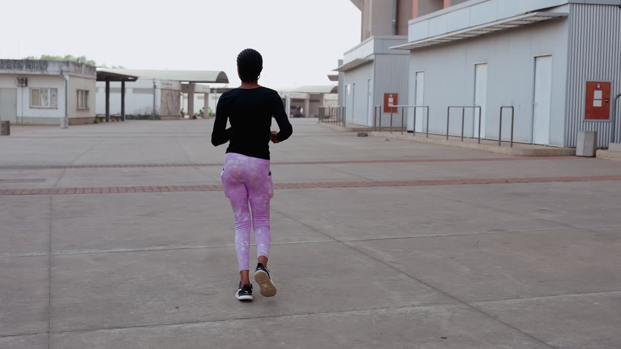 Persona che corre attraverso un cortile industriale vuoto, corridore urbano che si allena da solo all'alba indossando un top nero e leggings viola, facendo jogging su un'ampia spianata di cemento tra i magazzini, senso di determinazione.