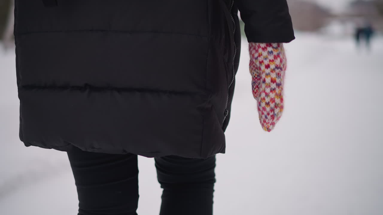 Lower body of Female wearing black winter coat walking on snowy path with colorful knitted mittens visible close up back view cold season outdoor scene blurred people in distance