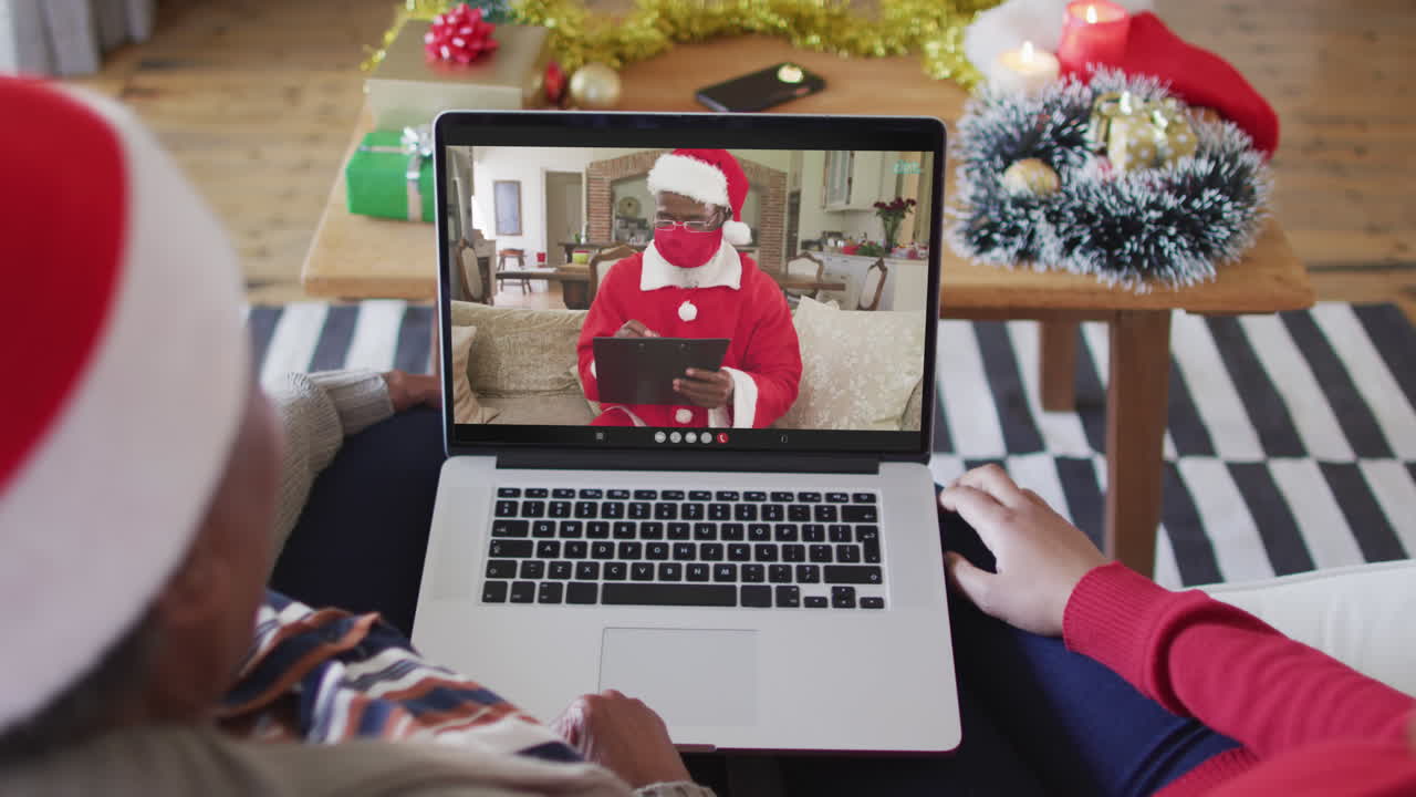 madre y hija afroamericanas usando una computadora portátil para una videollamada de navidad con santa en la pantalla