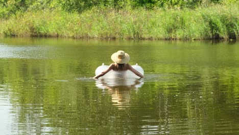 woman in swimsuit and straw hat is floating with an inflatable circle in lake river pond. girl rests on swimming ring in a pool with blue water. concept of travel, vacation, weekends, holidays, relaxing on nature, rest, relaxation