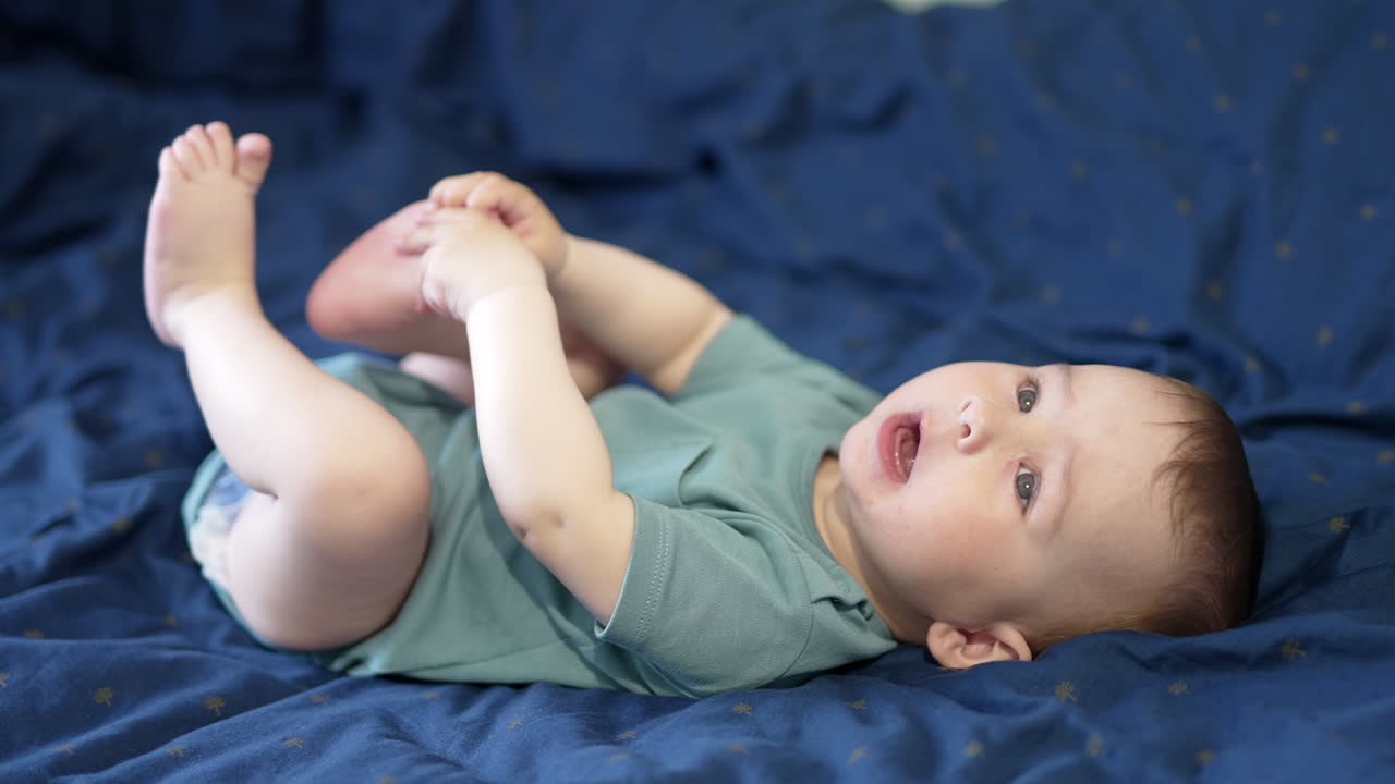 Pretty little kid actively moving while lying on the bed. Adorable baby boy interested in his legs pulling them closer. Blue background.