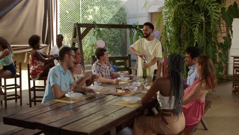 A diverse group of friends gathered around an outdoor table, enjoying food and wine at a casual party