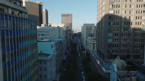 Backwards-fly-above-wide-boulevard-surrounded-by-tall-buildings-in-city-centre.-Cape-Town,-South-Africa