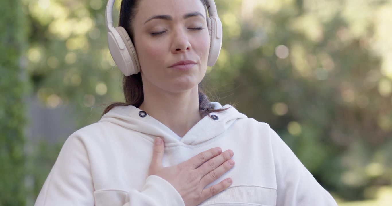 Donna adulta con le mani sul petto e sull'addome, che medita in giardino indossando una felpa leggera con cappuccio e delle cuffie.