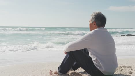 back view of hispanic senior man sitting on beach at sunset