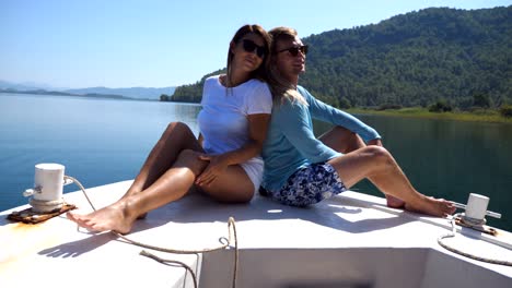 young pair sitting back to back and posing on bow of boat at sunny day. happy couple in love spending time together on deck of ship and enjoying summer travel. concept of vacation or holiday. close up