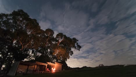 a zoomed in time lapse of a man having a camp fire by a shed at night under some gum trees with clouds rolling past and the night time stars in the background