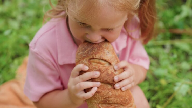 white toddler girl grimacing after bite, rubbing forehead and wiping crumbs from cheek, pausing in thought then offering hesitant smile, contemplative moment under trees during outdoor snack, candid