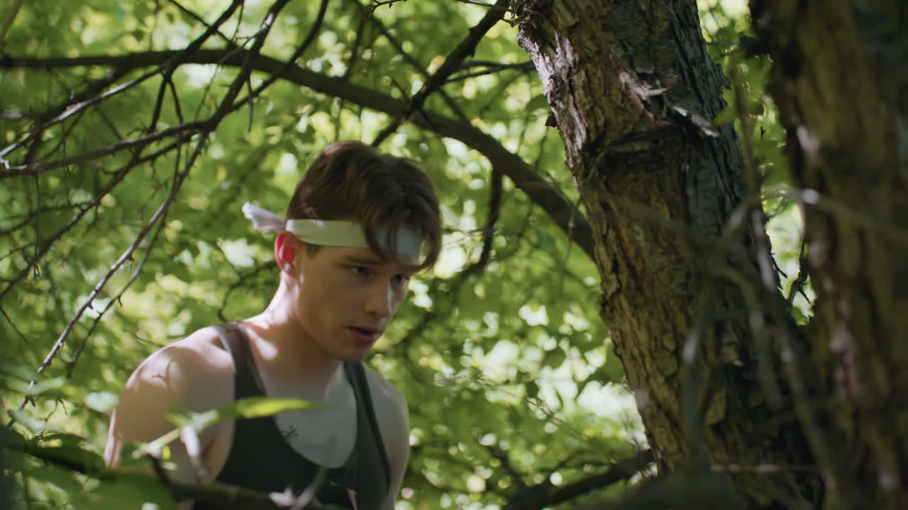 soldier wearing headband and gloves repeatedly punches rough tree bark under dense canopy, sweat and fatigue visible on determined face as he trains endurance and strength in secluded woodland