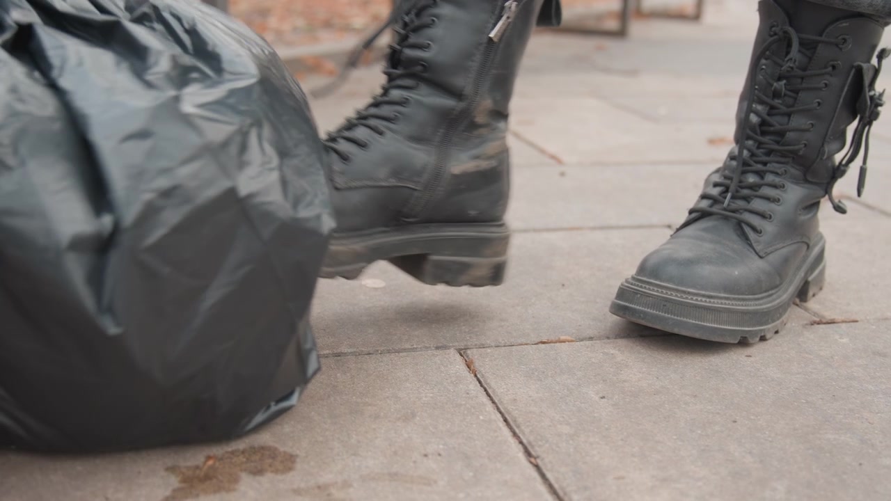 Black trash bag on sidewalk closeup showing heavy boots nearby, plastic sack crumpled on paved pavement, gritty urban atmosphere with scattered litter, low angle reveal of approaching footsteps