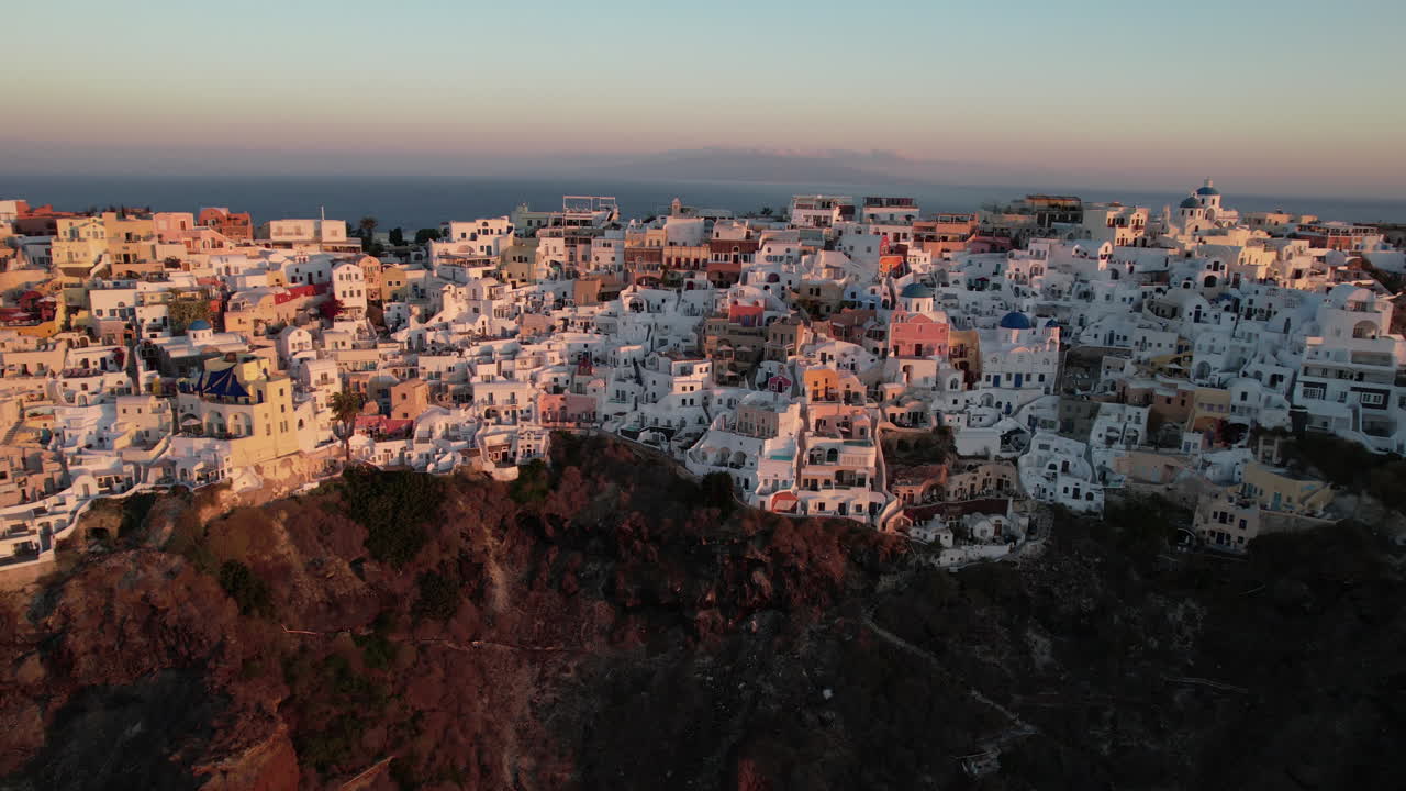 vista aérea de un pueblo en rocas rojas y acantilados de la isla de santorini, grecia, al atardecer, tomada por un avión no tripulado
