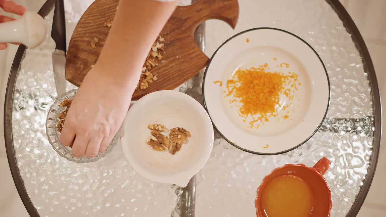 Topdown kitchen view with pumpkin zest, hands arranging walnuts and zest on plate, warm light on glass table, mug of juice nearby, intimate cozy atmosphere, methodical recipe staging