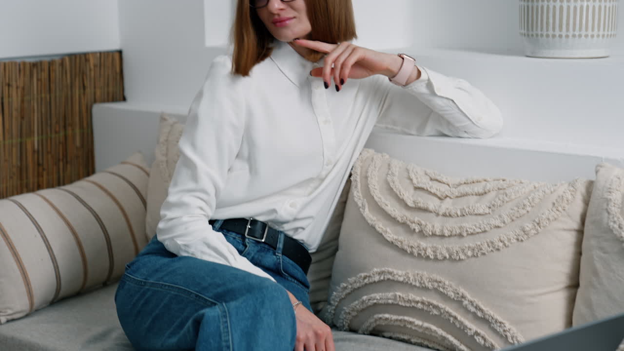 Woman wearing white shirt and jeans sits on sofa. Opened laptop is next to the lady on the sofa.