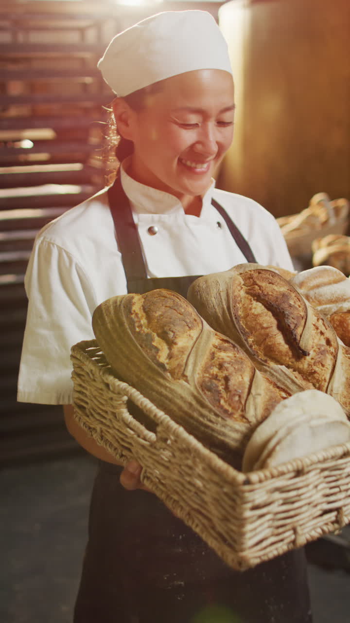 Vertical video of portrait of happy asian female baker with bread in bakery