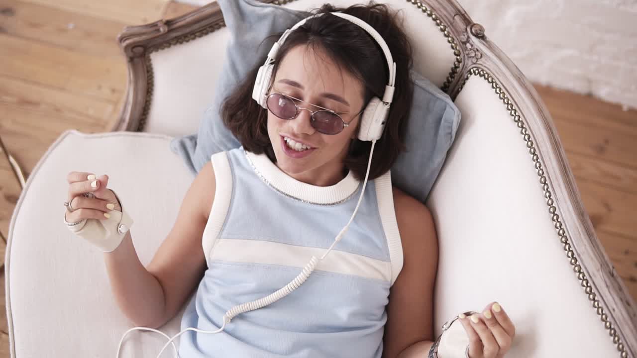 Stylish hipster girl in her 20's wearing headphones, glasses and punk leather biker-gloves, lying on the couch, singing and dancing to the music