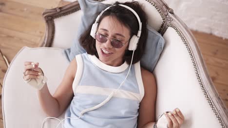 Stylish-hipster-girl-in-her-20's-wearing-headphones,-glasses-and-punk-leather-biker-gloves,-lying-on-the-couch,-singing-and-dancing-to-the-music