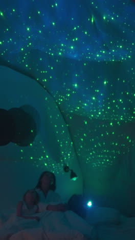 la madre y los niños se sientan en la cama mirando y estudiando el cielo estrellado multicolor proyectado en el techo blanco por el proyector. fuente de luz tenue en premisa semi-oscura