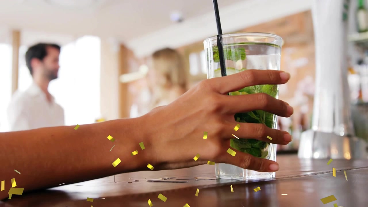 bartender picking mojito glass in bar, lifting toward camera to serve couple with gold confetti