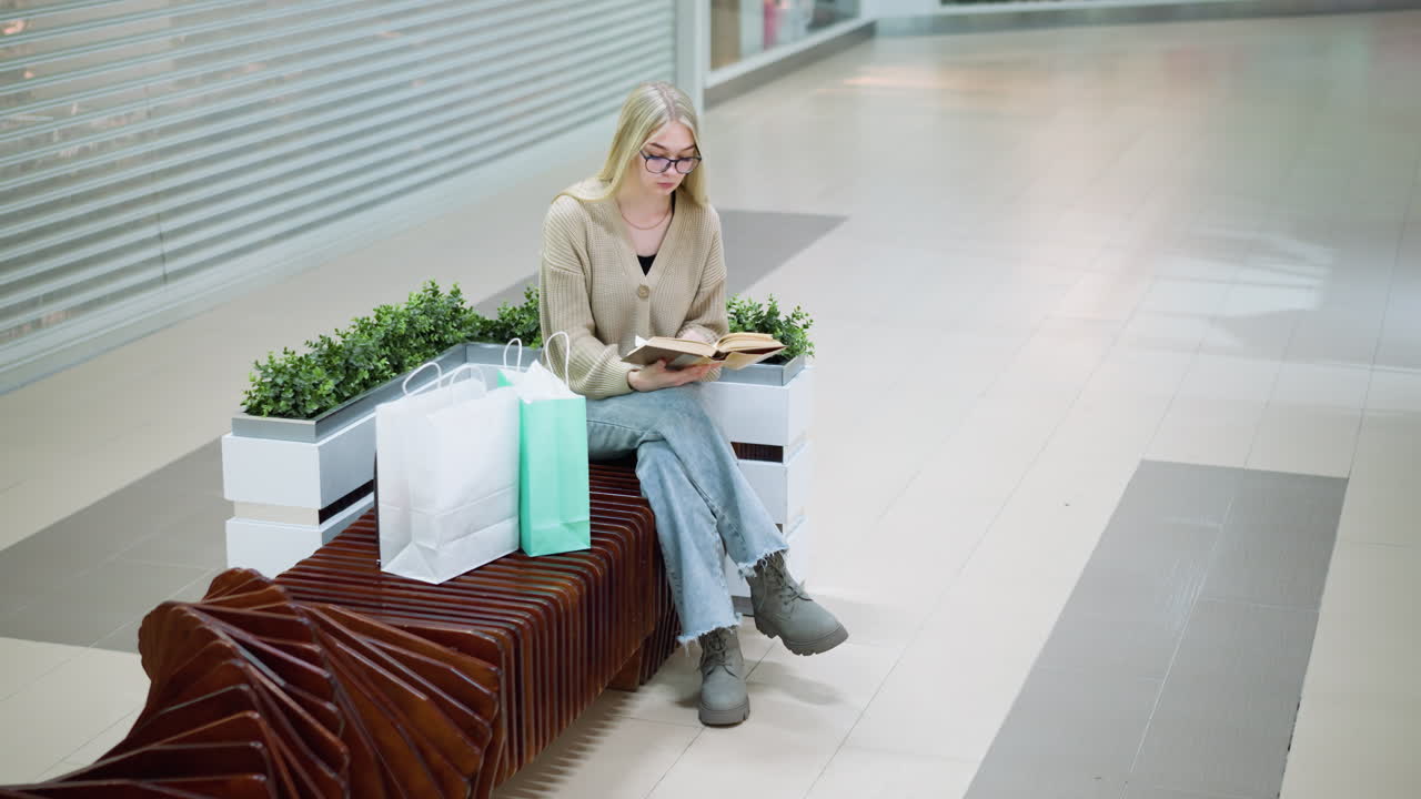 Ung dame sidder i et lyst indkøbscenter med benene over kors og læser en bog, med indkøbsposer placeret ved siden af hende på bænken, en afslappet shoppingoplevelse med afslappet stil.