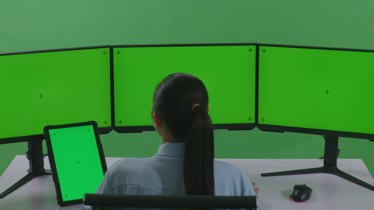 태블릿과 여러 컴퓨터 모니터로 작업하는 아시아 여성의 뒷면은 녹색 스크린 스튜디오에서 녹색 화면 모형을 가지고 있습니다.