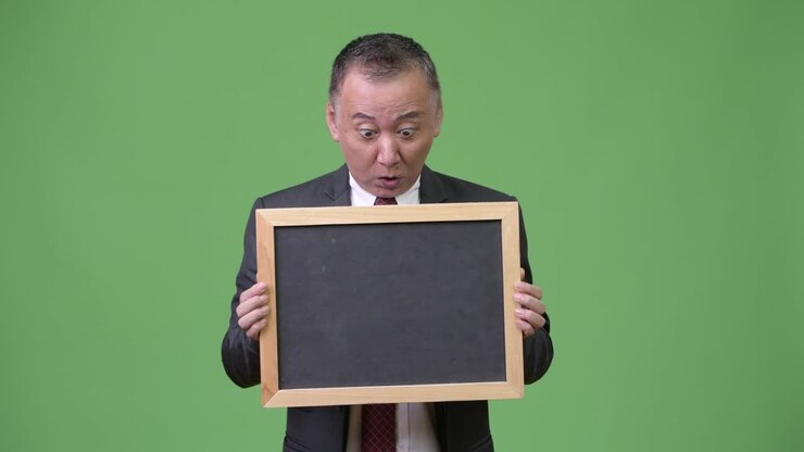 Mature Japanese businessman holding blackboard against green background