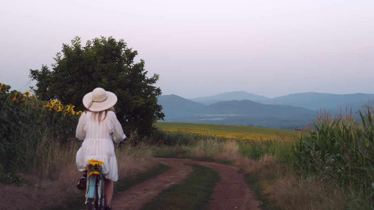 드레스를 입은 소녀가 평온한 시골에서 골든 아워에 천천히 자전거를 타고 있습니다.