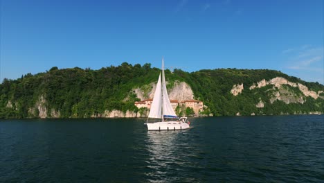 Aerial-View-Of-serene-scene-of-a-white-sailboat-navigating-calm,-blue-waters-near-a-rocky,-lush-green-cliffside-on-a-clear,-sunny-day-under-a-vibrant-blue-sky