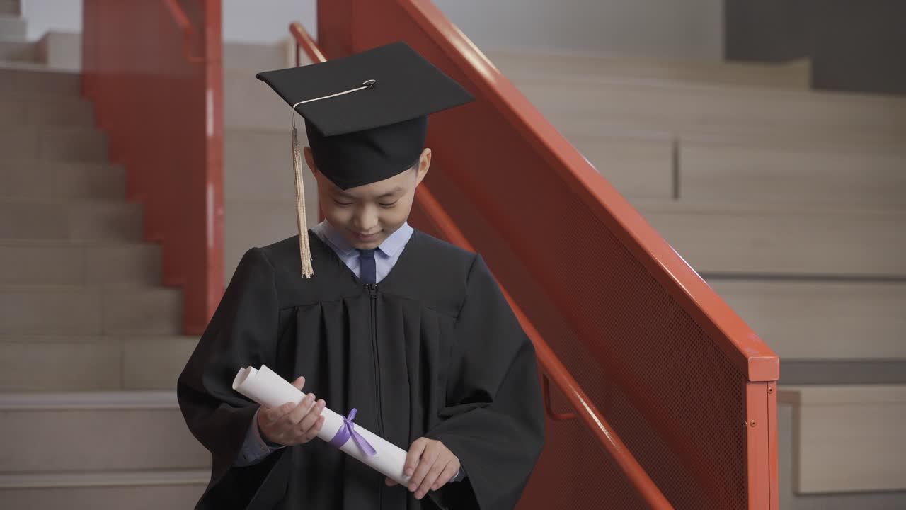 feliz estudiante asiático de preescolar con toga y birrete sosteniendo un diploma y celebrando su graduación