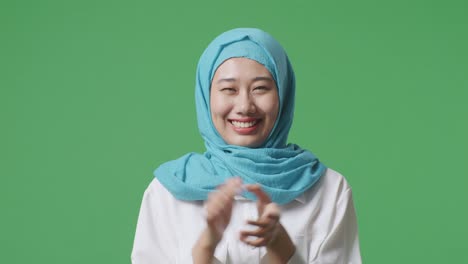 녹색 스크린 배경 스튜디오에 서 있는 아시아 무슬림 여성이 미소 짓고 손을 치고 있습니다.