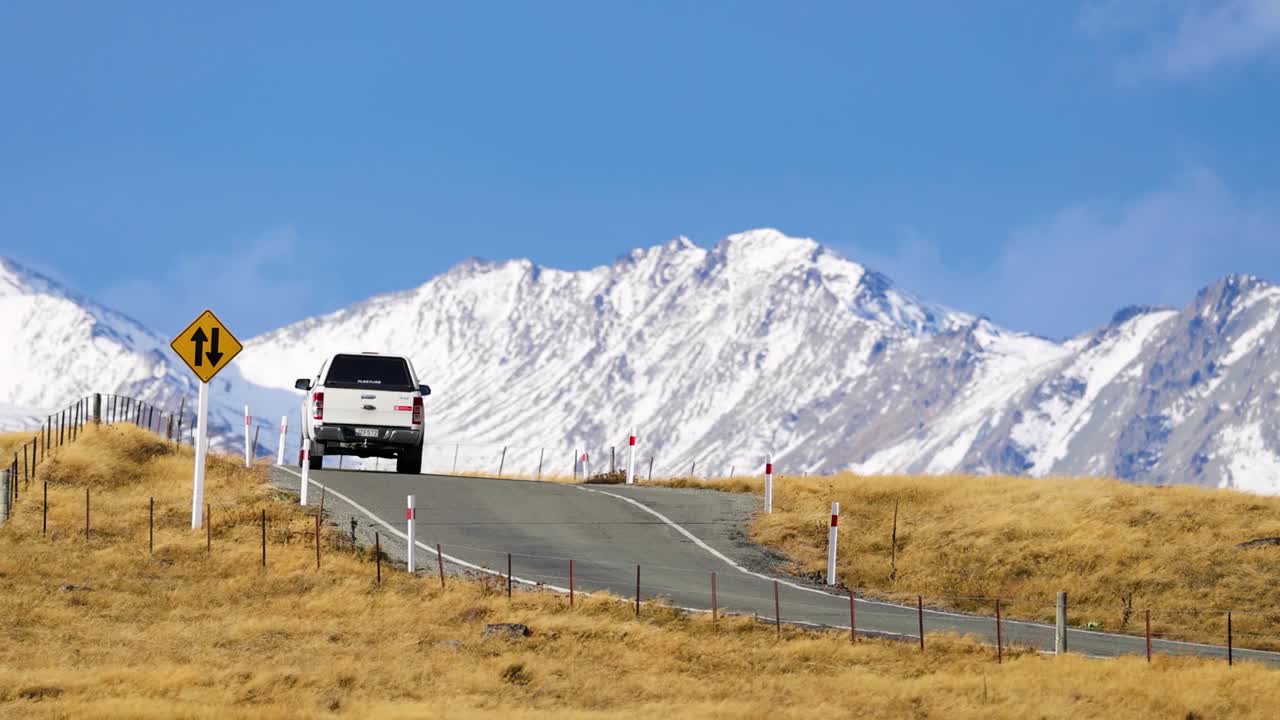 A white SUV travels along a winding rural road through golden grasses toward snow-covered mountains under clear blue skies, captured in steady daylight