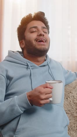 handsome smiling indian man drinking warm coffee while sitting on couch in living room at home