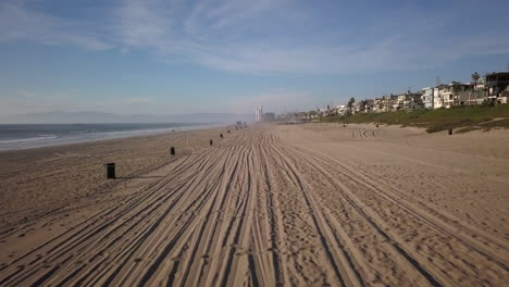 aerial-shot-at-golden-hour,-going-forward-above-the-sand-of-manhattan-beach,-with-car-prints-on-the-sand