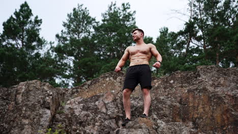 caucasian muscular man in black shorts stand high on the rock. sportsman demonstrates his athletic torso. low angle view.