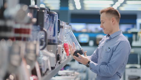 in the appliances store, a man in a shirt chooses a blender to buy by viewing and holding the device.