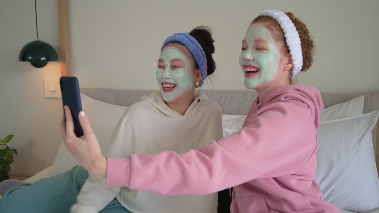 Forskellige veninder sidder på sengen med grønne masker, og kvinden til højre holder en telefon og tager spa-selfies