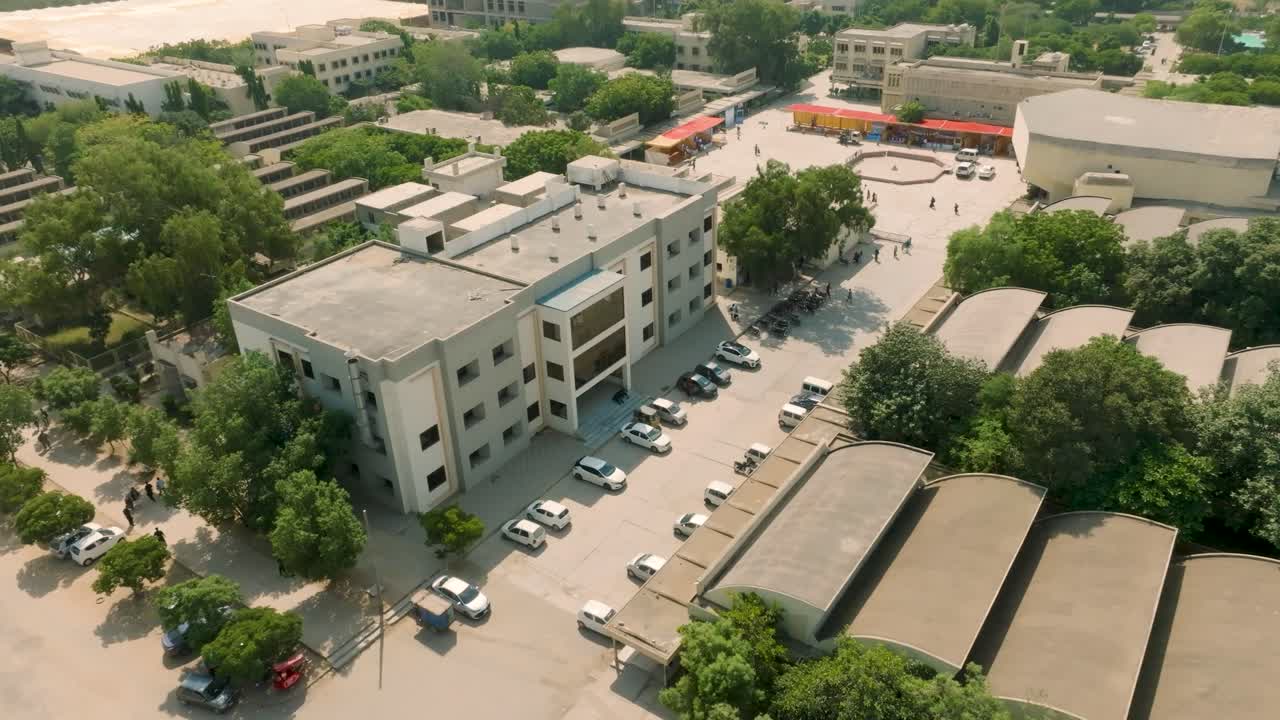 Aerial orbit view of NED University of Engineering and Technology campus parking lot in Karachi, featuring parked cars, students, and campus buildings under a sunny sky