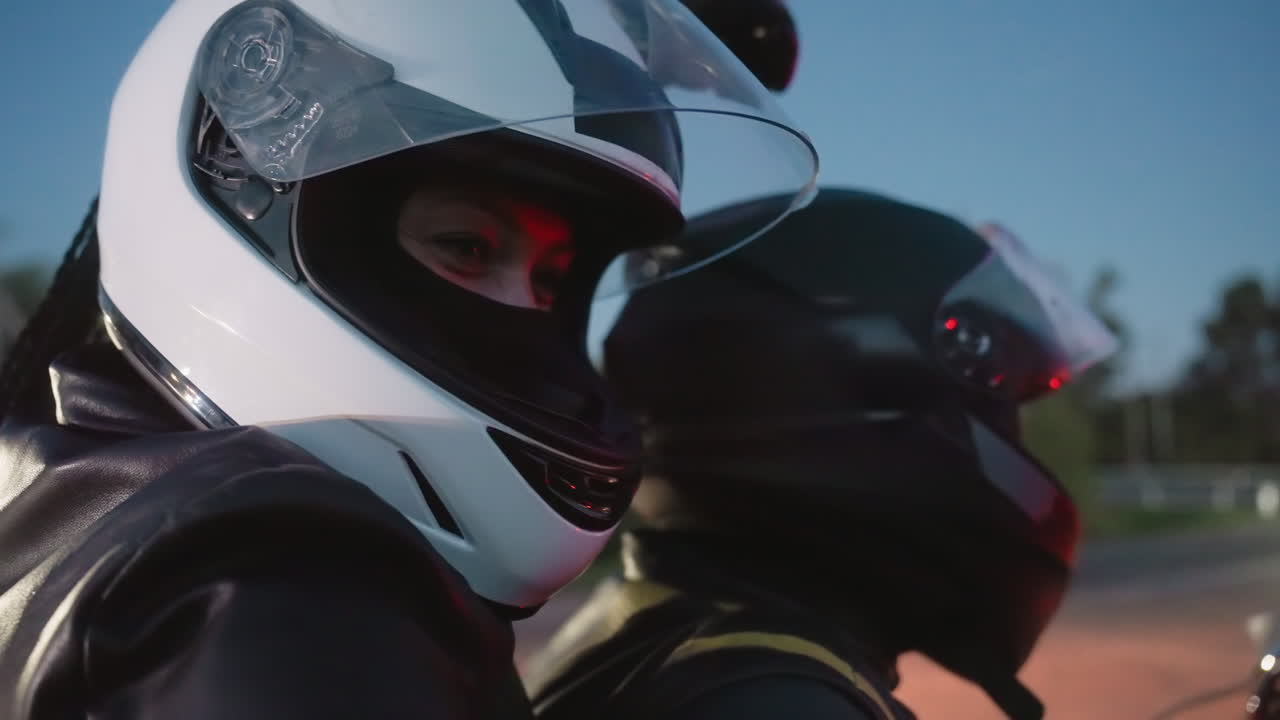 Lady wearing helmet takes picture with man on motorcycle as car passes by in background, evening ride atmosphere captured with glowing light reflections and candid expressions of couple in focus