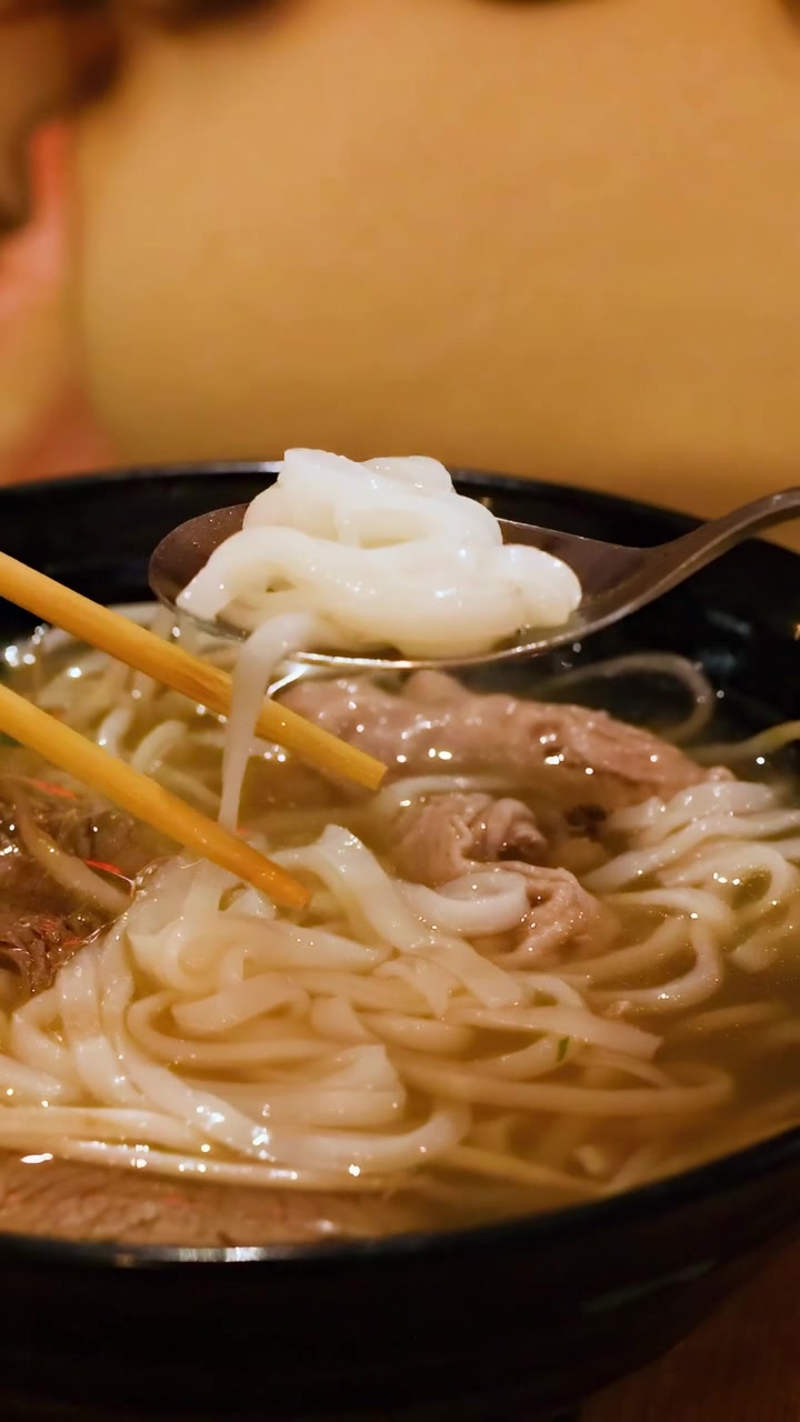 Cận cảnh sợi phở đang được gắp bằng đũa và thìa, làm nổi bật kết cấu và sự ấm áp trong một không gian ấm cúng
