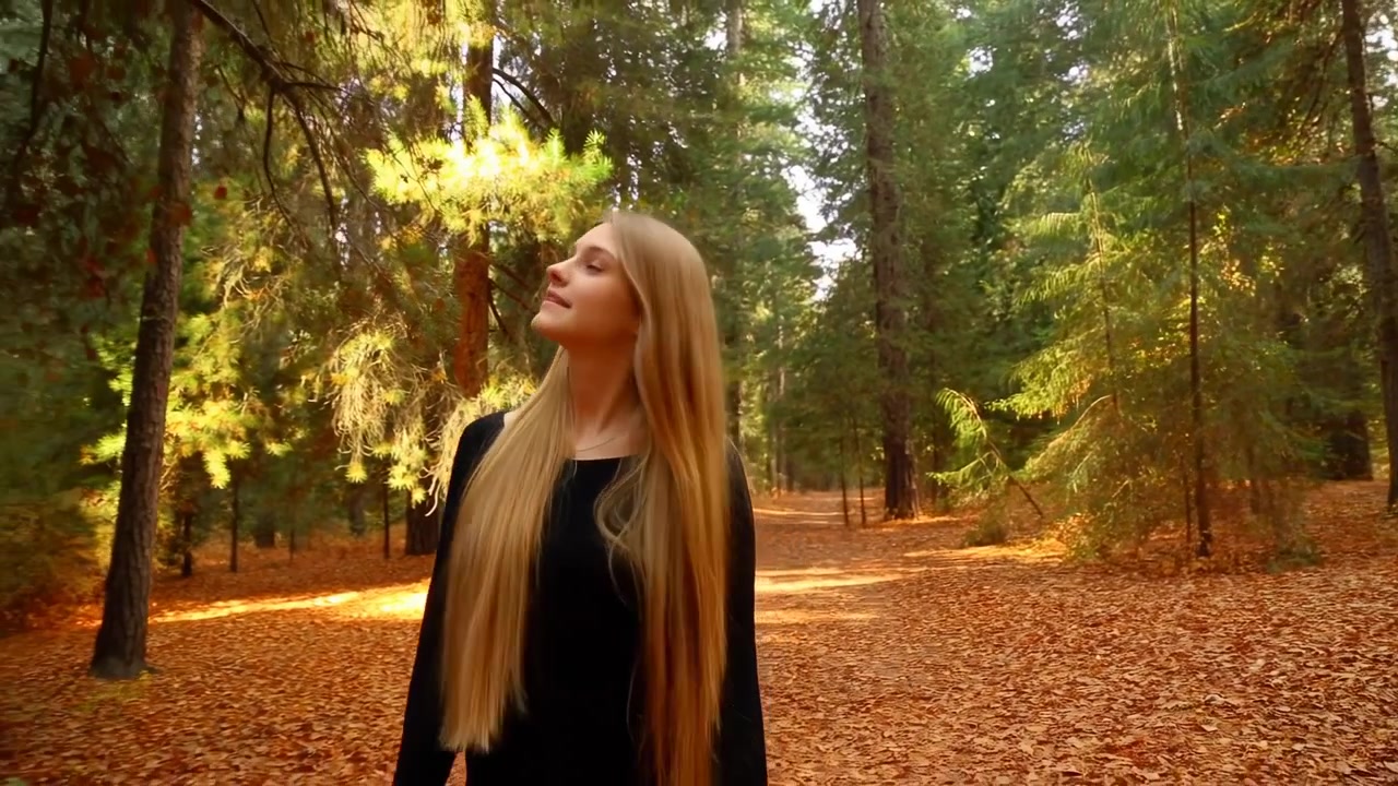 Giovane donna con lunghi capelli biondi si trova su un sentiero forestale coperto di foglie, con lo sguardo rivolto in alto e a sinistra con calma curiosità, la luce soffusa del sole filtra tra gli alberi, creando un'atmosfera naturale boschiva.
