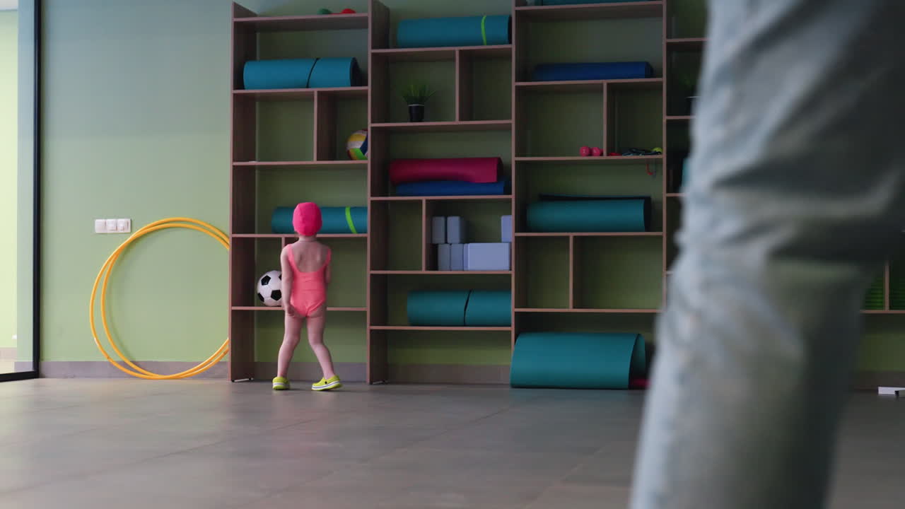 Attractive view of shelf with foam materials, ball, clock, small girl in swimming pants walking and touching items, curious gaze, warm indoor light, playful mood, tidy arrangement near pool