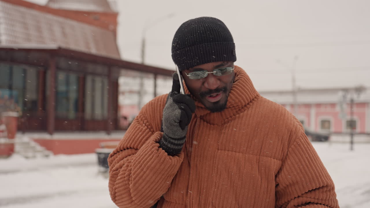 Uomo di colore che sorride mentre telefona, che indossa un cappotto di maglia arancione e un cappello nero, piazza urbana con neve che cade, tono amichevole, persona di piccolo imprenditore, gesti animati, clip di vita spontanea, caldo
