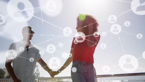 network of profile icons against mixed race couple holding hands walking on the beach